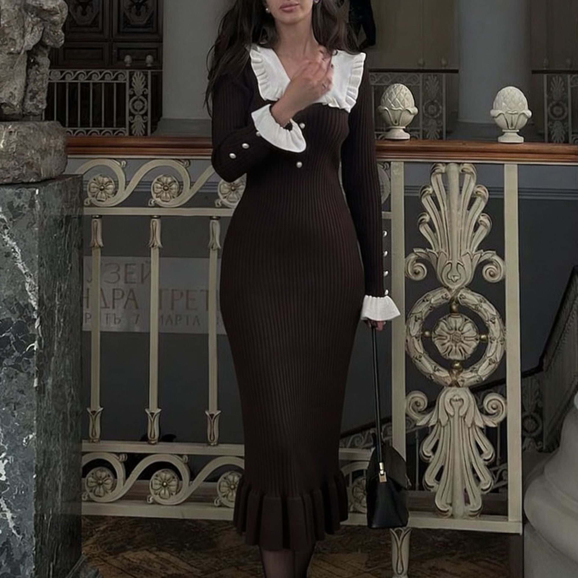 Woman in a dark brown dress with white ruffled collar standing in an ornate interior setting.