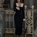 Woman in a black dress with white ruffled collar standing in an ornate interior setting.