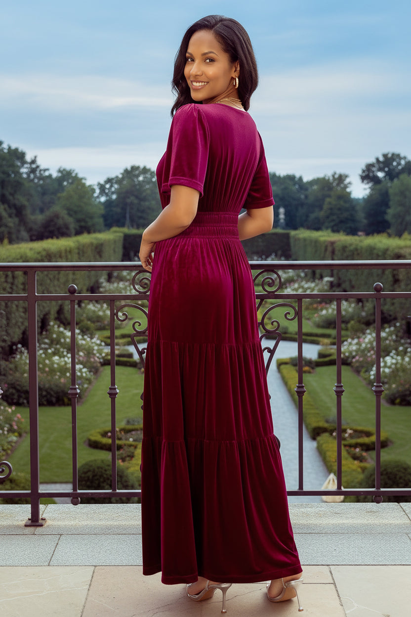 A woman wearing a deep magenta velvet maxi dress with short puff sleeves, shown from the back in an outdoor setting with a stone pillar and wrought iron gate.